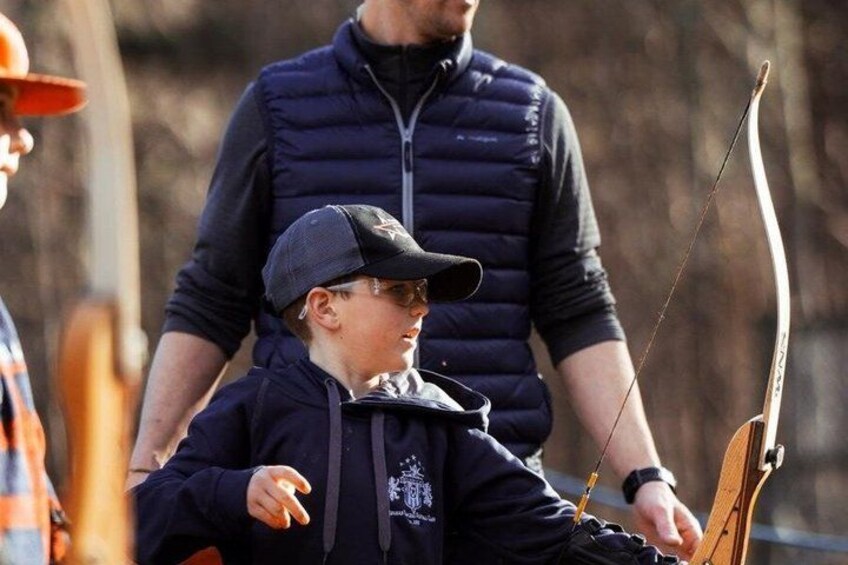 A person in a dark jacket and cap holding a bow and arrow, with another person standing behind. 5