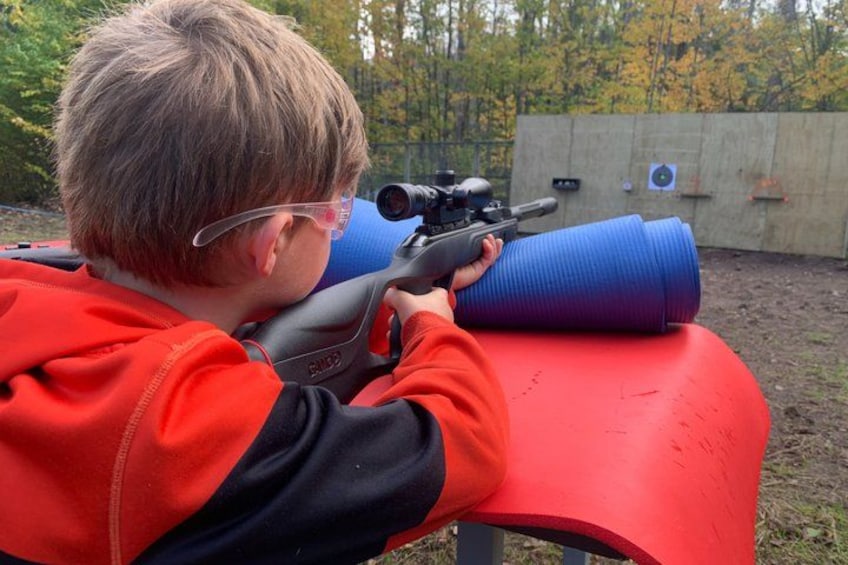 A person aiming a rifle with a scope, sitting on a red bench outdoors with trees and a fence in the background. 2