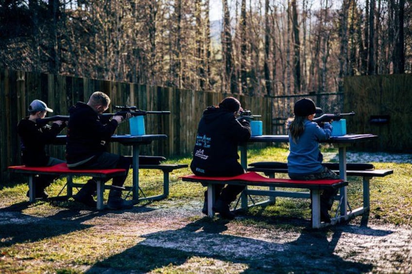 People aiming rifles at targets in an outdoor setting with benches and a wooden fence in the background. 5