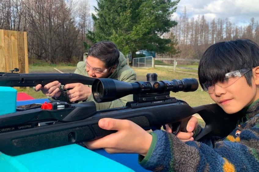 Two individuals aiming rifles with scopes, a blue tarp on the ground, and a wooden fence in the background. 1