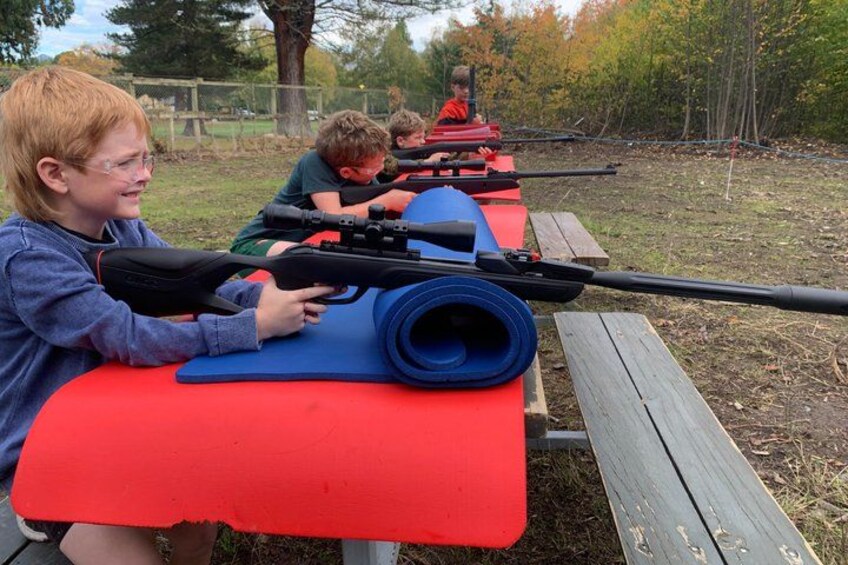 Children aiming rifles at targets on a red table in an outdoor setting. 4