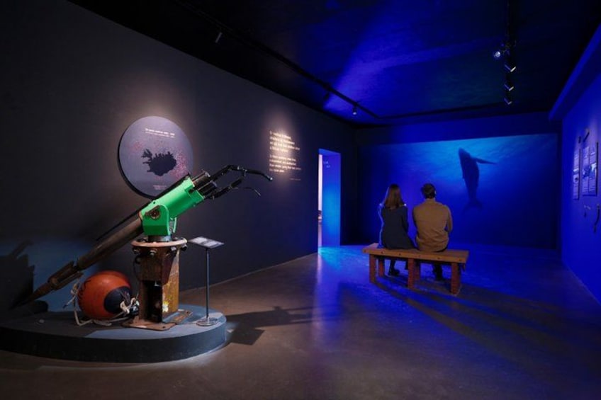 A museum exhibit with a robotic arm, a circular display, and a bench with two people observing a large screen. 3