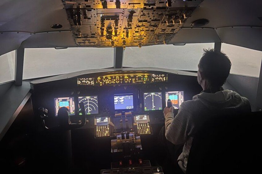 A cockpit with multiple control panels and screens, a person standing at the controls, and a view of the runway outside. 5