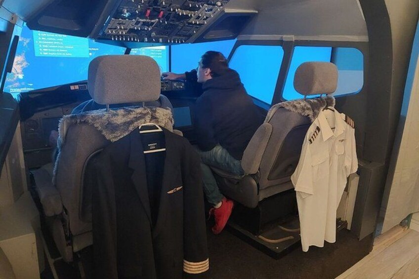 A cockpit with two pilots, one in a black jacket and the other in a white jacket, with a control panel and a screen displaying flight information. 3