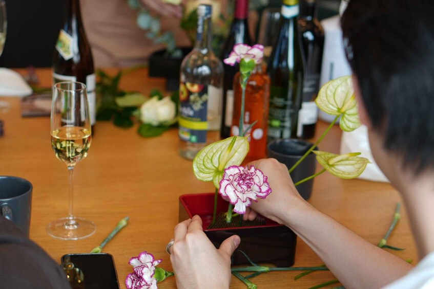 En person ordner blomster i en rød kasse på et bord med vinflasker og glas. 1