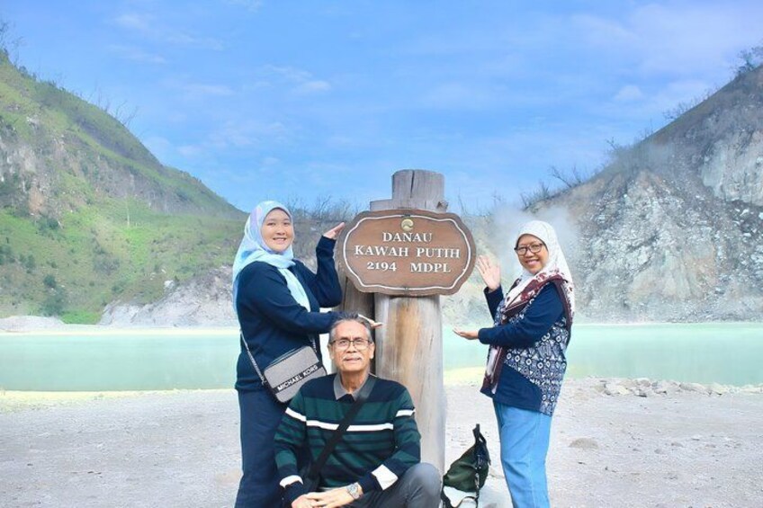 Three people standing in front of a wooden sign with text, a body of water, and a mountainous landscape in the background. 3