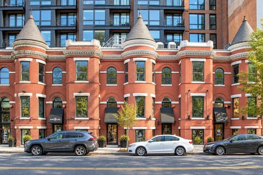 A row of red brick buildings with arched windows and towers, parked cars in front. 2