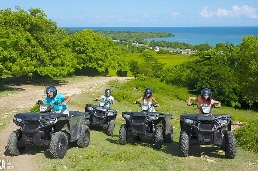 Four people on ATVs on a grassy hill with a view of the ocean and greenery in the background. 5