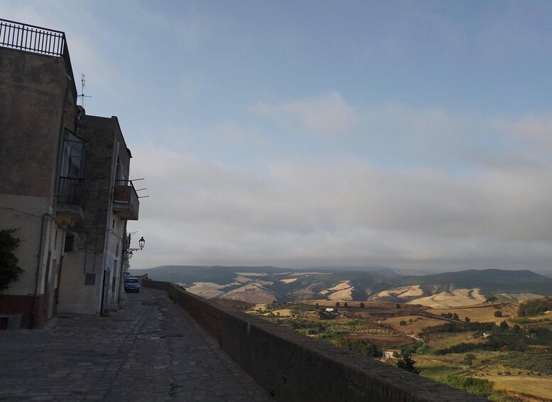 Une rue pavée de pierres, bordée de bâtiments d’un côté et offrant, au loin, une vue sur des collines ondulantes. 5
