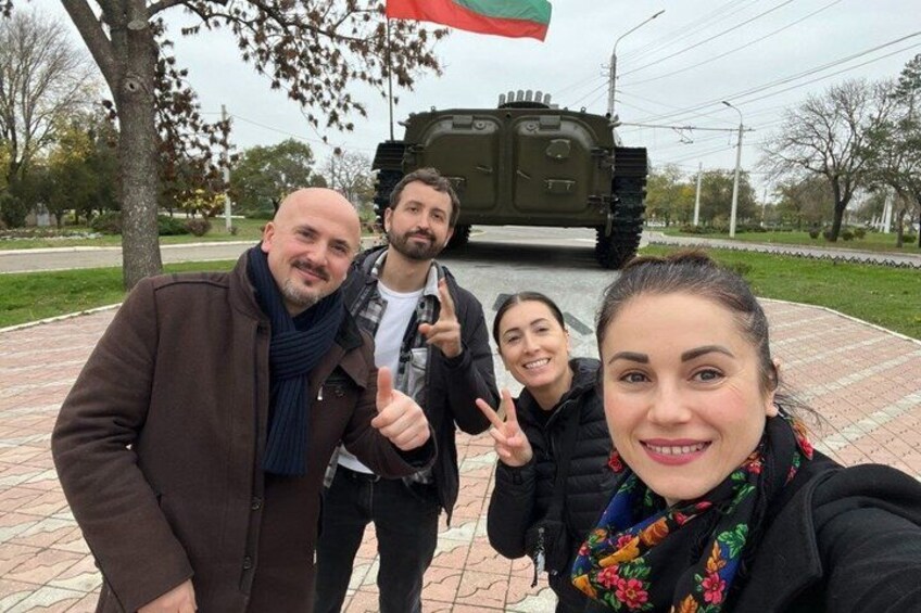 A group of people posing with a military vehicle in a paved area with trees and a grassy field in the background. 1