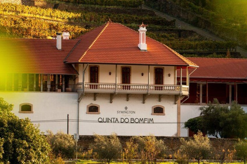 A white building with a red roof, named 'QUINTA DO BOMEIM,' surrounded by greenery and hills. 1