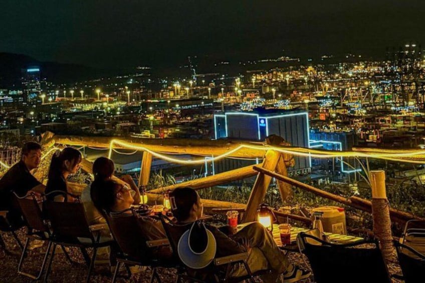 A group of people sitting on chairs at night, overlooking a cityscape with illuminated buildings. 4