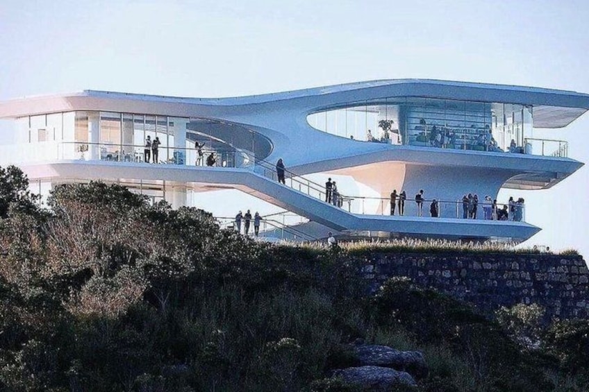 A modern architectural structure with a curved design, situated on a grassy hill with a stone wall in the foreground. 3
