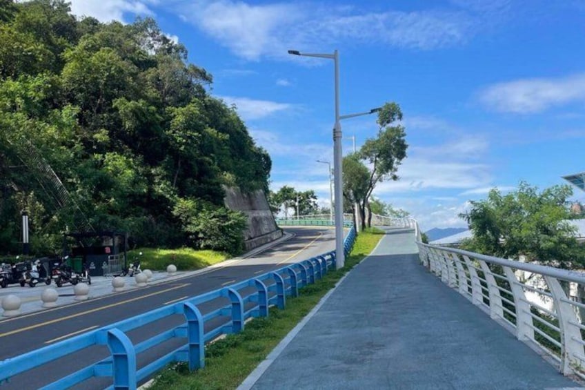 A pedestrian pathway alongside a road with a blue guardrail, trees, and a clear sky. 1