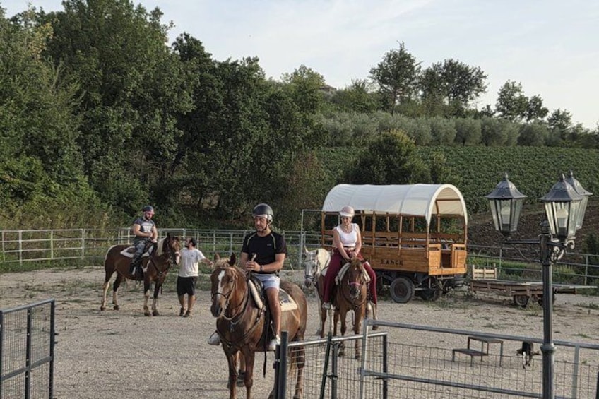A group of people riding horses in an outdoor arena with a covered wagon and lampposts in the background. 1