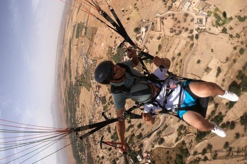 A person paragliding over a desert landscape with sparse vegetation and distant buildings. 4