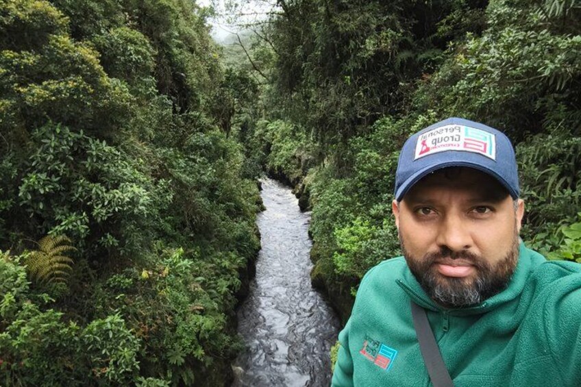 A person in a green jacket and blue cap standing in a lush green forest with a river in the background. 3