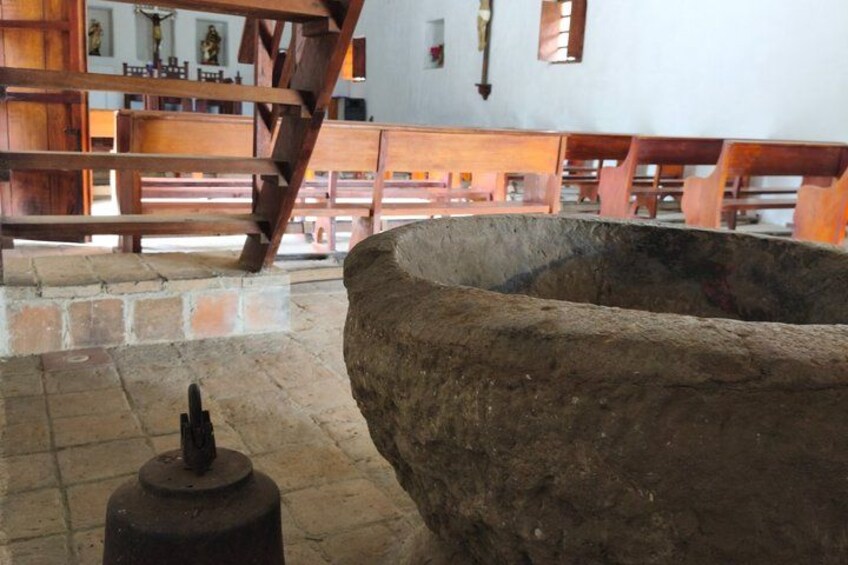 A church interior with wooden pews, a large stone basin, and a metal object on the floor. 4