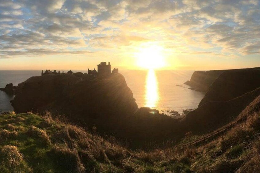 A coastal landscape with cliffs, a castle, and the sun setting over the horizon. 5
