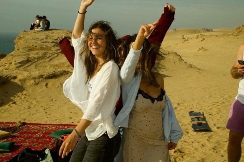 Two people on a sandy beach with a red blanket and a green bag. 1