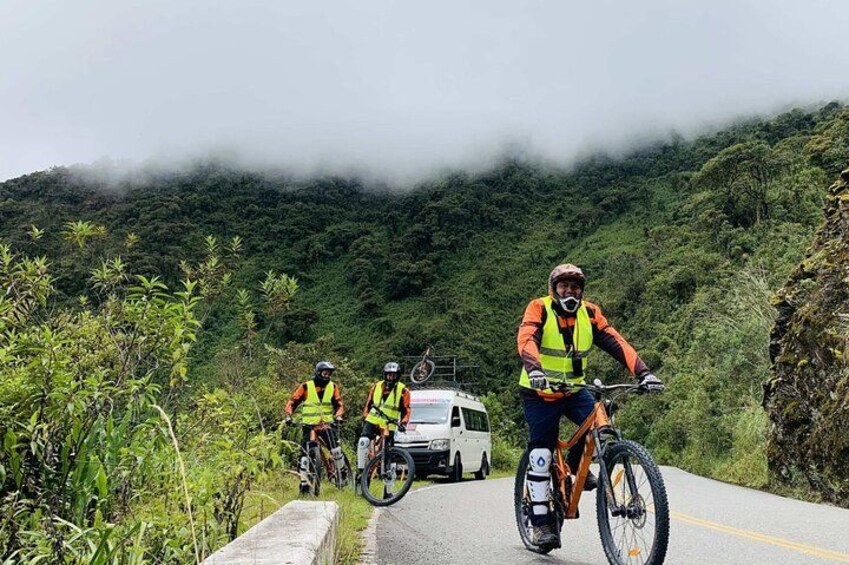Three cyclists on a mountain road with a white vehicle in the background. 1