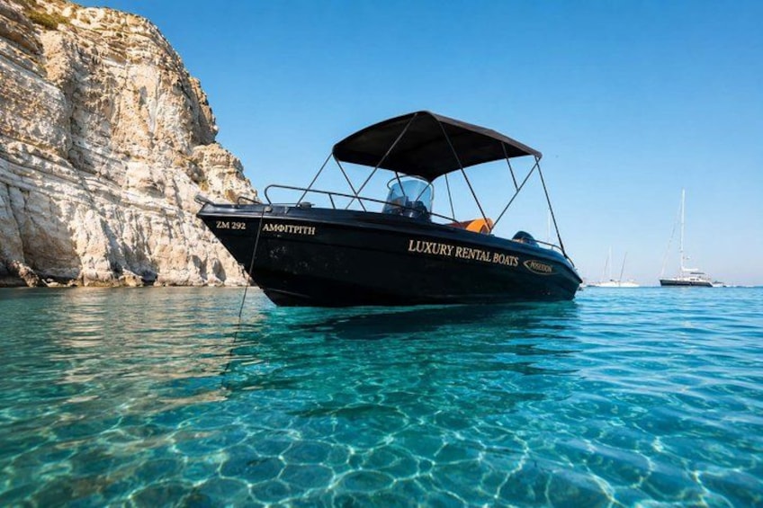 A black boat named 'Luxury Rental Boats' with a canopy, anchored near a rocky cliff and clear blue water. 1
