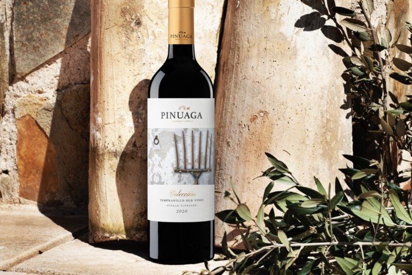 A bottle of Pinuaga wine with a label featuring a fork and knife, set against a stone wall with greenery. 3