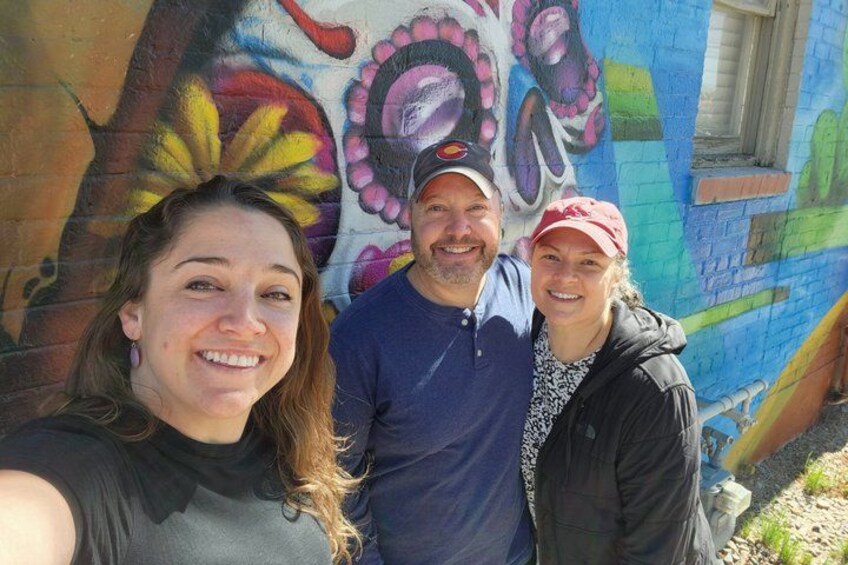 Three people standing in front of a colorful mural on a brick wall. 4