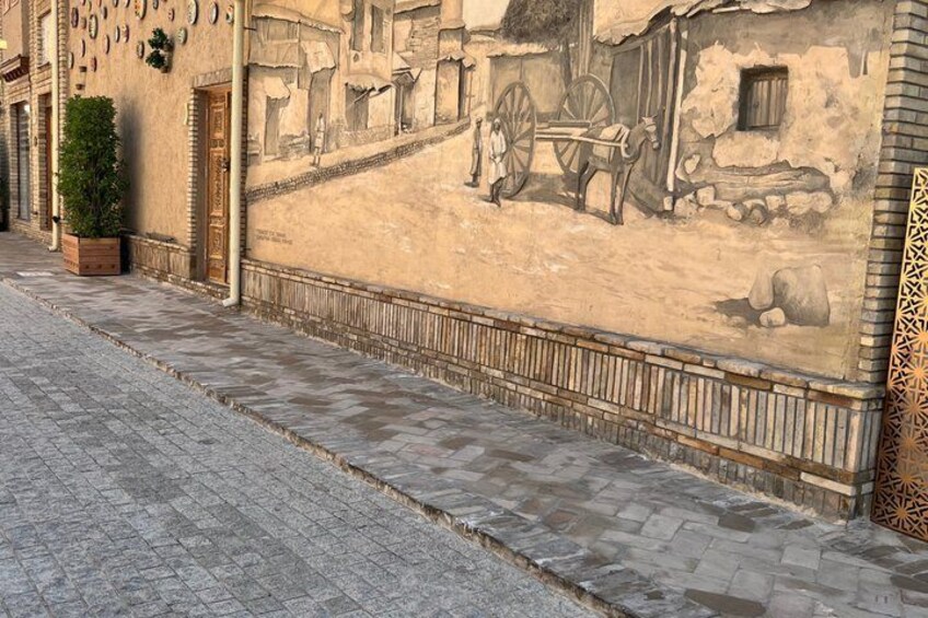 A cobblestone street with a mural depicting a rural scene, a wooden door, and a potted plant. 3