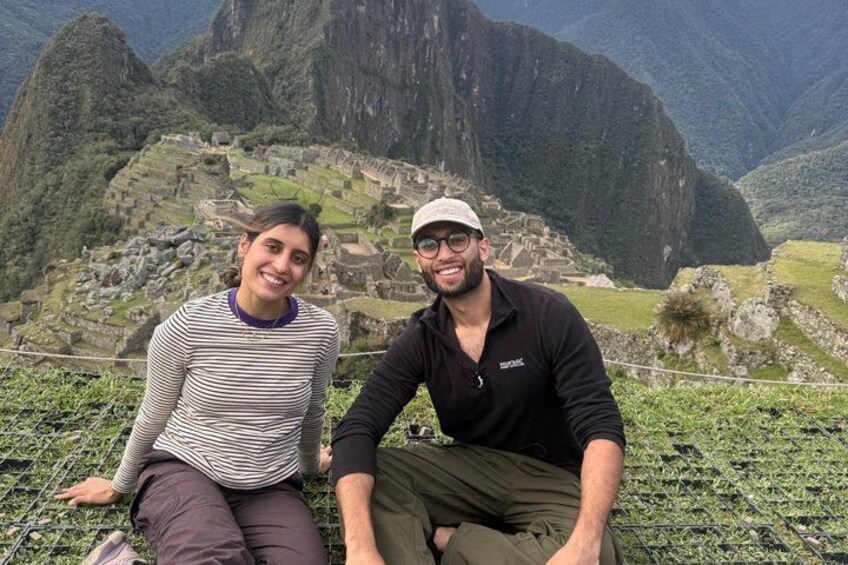 Two people sitting on a grassy area with Machu Picchu ruins in the background. 4