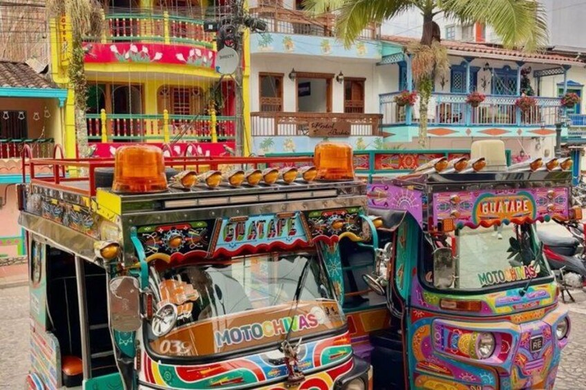 Colorful, decorated buses with 'GUATAPPE' and 'MOTO HVA' text, parked in a vibrant urban area. 5