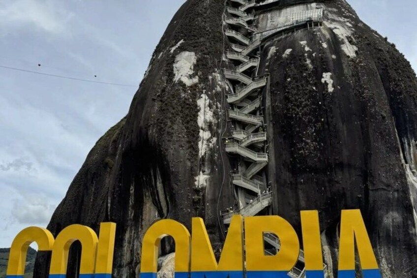 A large rock formation with a staircase leading up its side and the word 'Colombia' in yellow letters at the bottom. 4