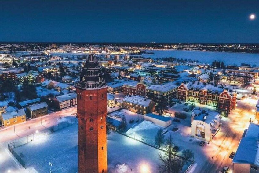 A snow-covered town with a prominent tower, illuminated buildings, and a river in the background. 1