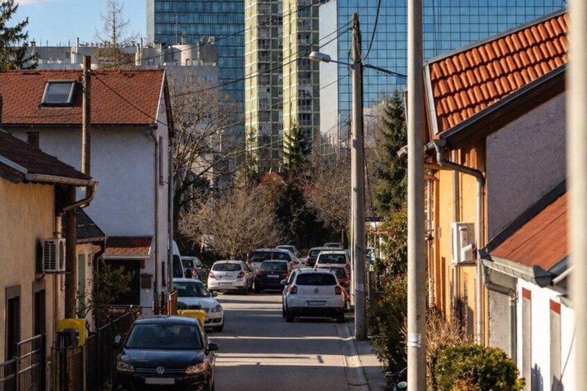 A narrow urban street lined with parked cars and residential buildings, with a tall modern building in the background. 2
