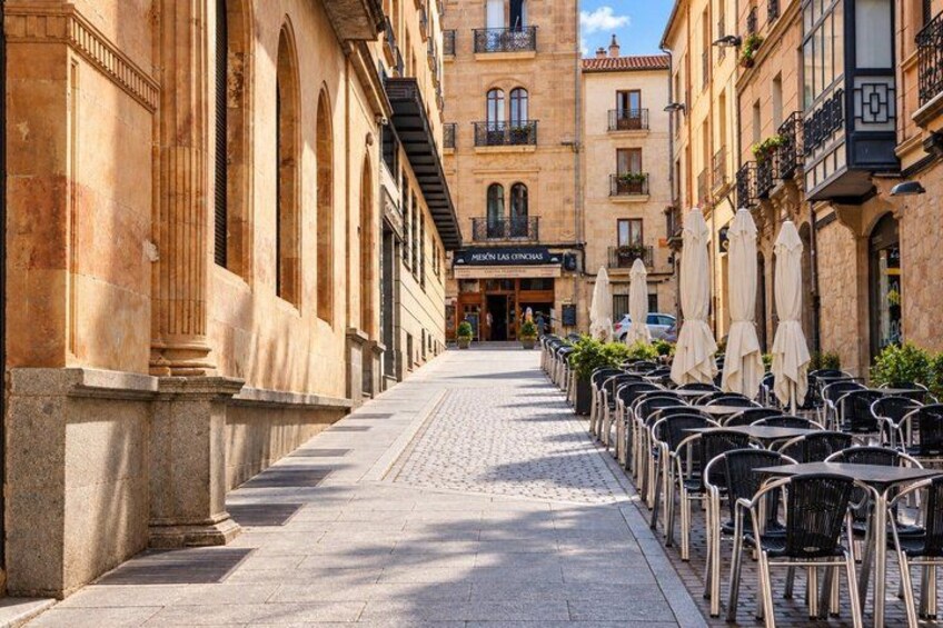 A cobblestone street lined with cafe tables and chairs, historic buildings with arched windows, and a clear blue sky. 5