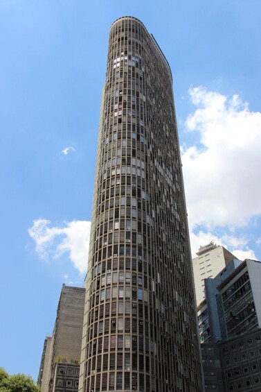 A tall, cylindrical skyscraper with a grid-like facade, surrounded by other buildings under a clear sky. 5