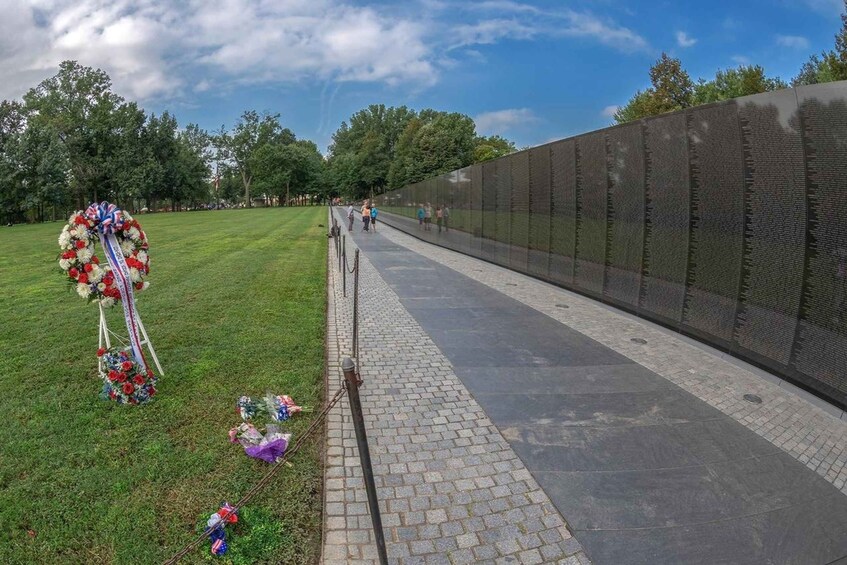Un muro conmemorativo con nombres grabados, un área verde con un ramillete y un sendero pavimentado. 5