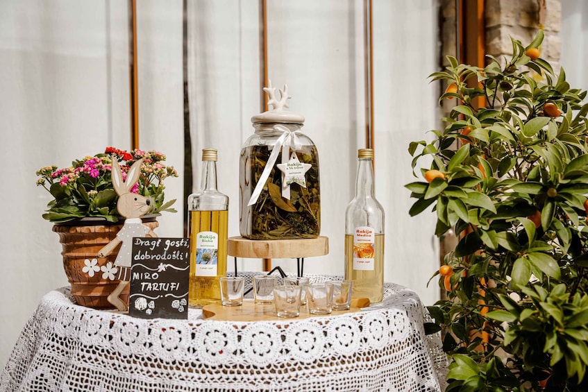 A table with a lace tablecloth, a wooden stand, a jar of leaves, two bottles of Rakija, a potted plant with orange fruits, and a decorative bunny. 3