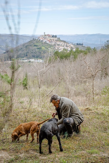 A person crouching on a hillside with two dogs, overlooking a village. 2