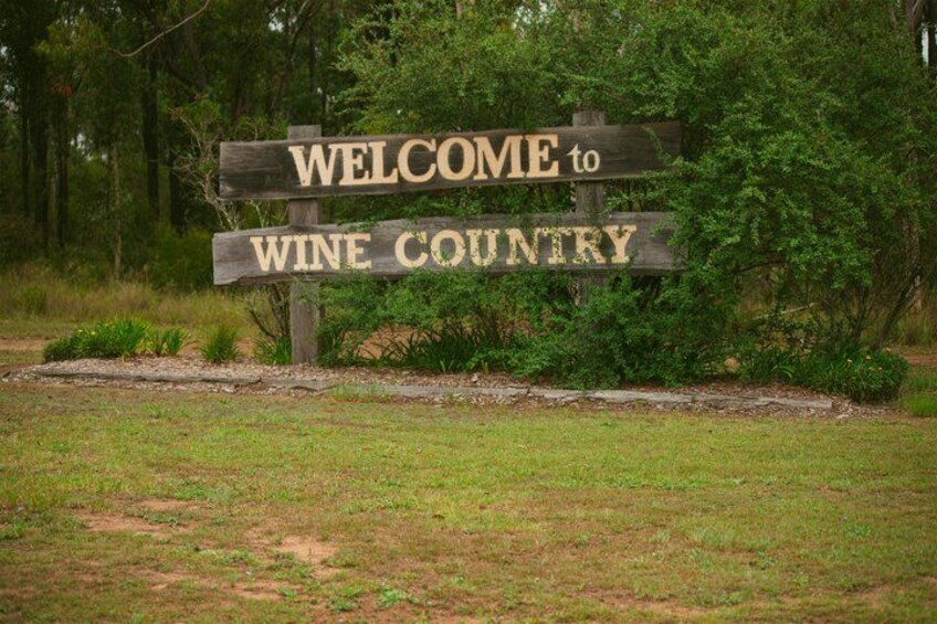 A wooden sign with the text 'WELCOME to WINE COUNTRY' set against a backdrop of trees and grass. 3