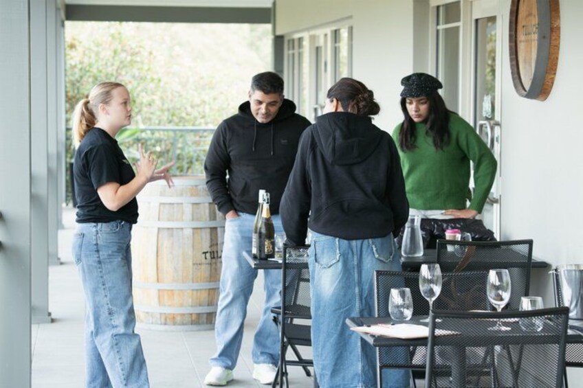 A group of people standing outside a building with a table and chairs, a barrel, and a bottle on the table. 2