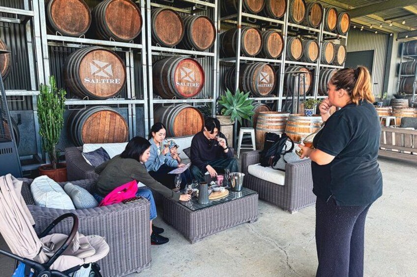 A group of people sitting on wicker furniture in an outdoor setting with wooden barrels in the background. 1