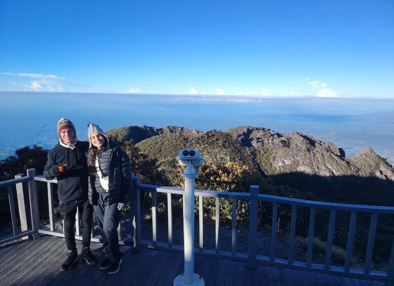 Two people standing on a wooden platform with a telescope, overlooking a mountainous landscape and a distant view of the ocean. 4