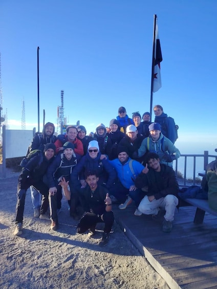 A group of people posing on a sandy area with a flag and communication towers in the background. 2