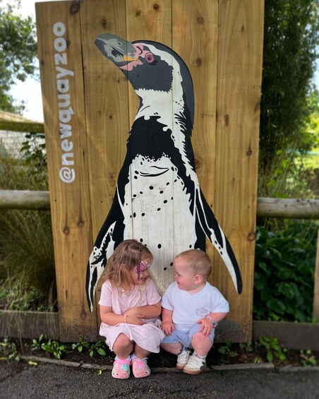 Two children sitting in front of a wooden sign with a painted bird and the text '@newquayzoo'. 5