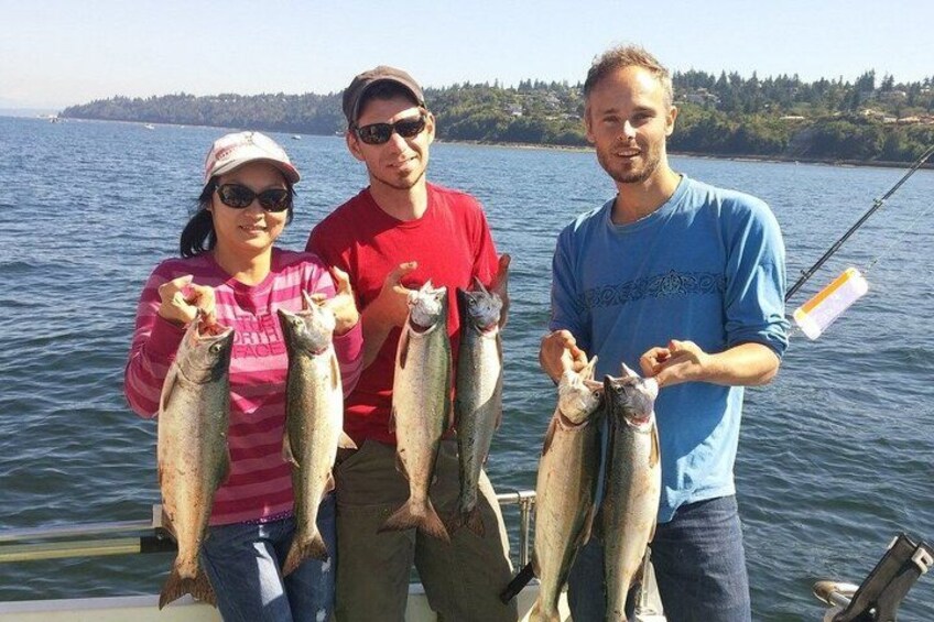 Three people holding fish on a boat with a lake and trees in the background. 4