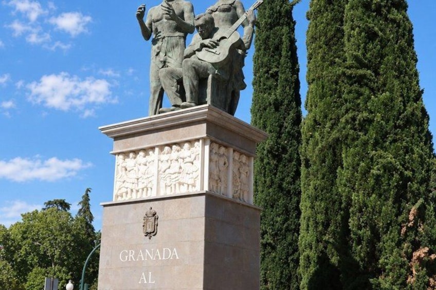 A statue on a pedestal with the inscription 'GRANADA AL' and a tall, narrow tree in the background. 5