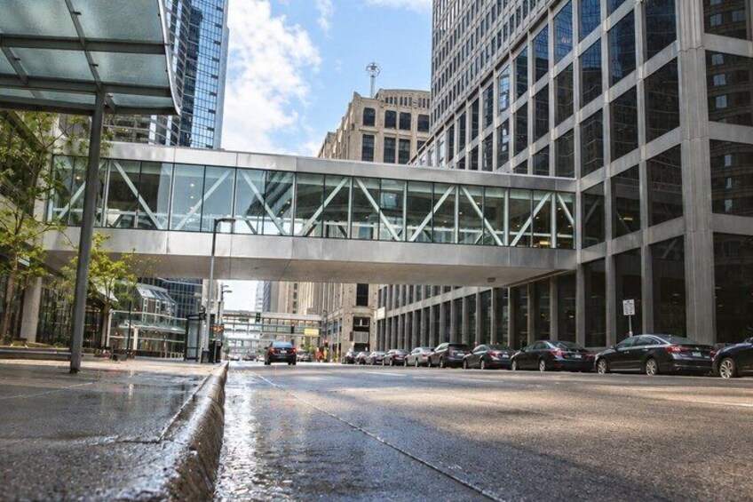A modern urban street with a glass pedestrian bridge, tall buildings, and parked cars. 5