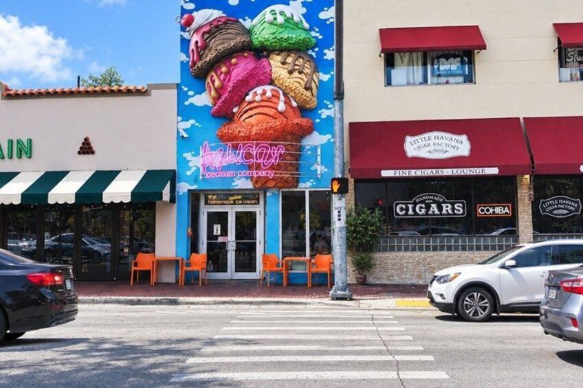 A street with a crosswalk, cars, and storefronts including a cigar lounge and a bakery. 5