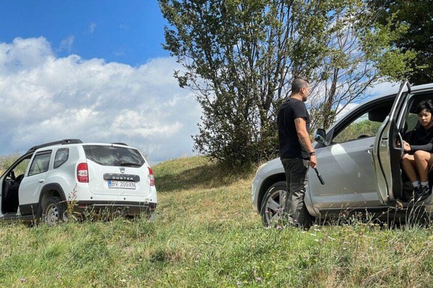 A white SUV parked on a grassy hill with its door open, a person standing beside it, and another person sitting inside. 4